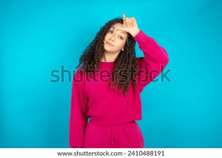 Beautiful Arab teen girl with curly hair wearing pink sweater making fun of people with fingers on forehead doing loser gesture mocking and insulting.