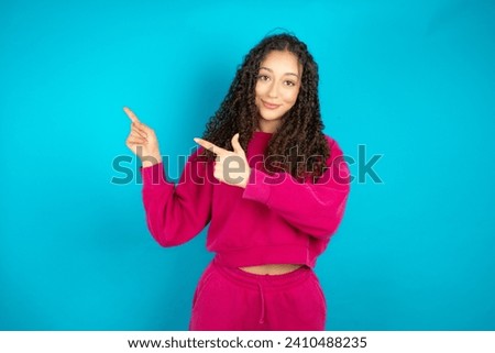 Beautiful Arab teen girl with curly hair wearing pink sweater indicating finger empty space showing best low prices, looking at the camera