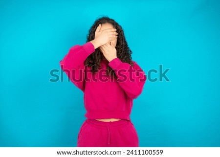 Beautiful Arab teen girl with curly hair wearing pink sweater Covering eyes and mouth with hands, surprised and shocked. Hiding emotions.