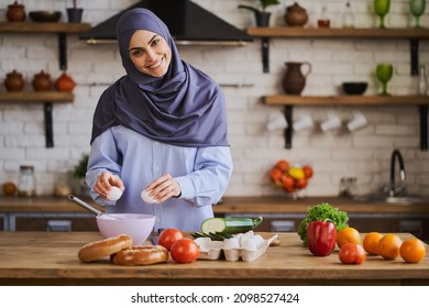 Beautiful Arabian housewife cooking a meal in her kitchen. Happy woman breaking the eggs into the bowl. Fruits, vegetables and bakery on her table. - Shutterstock ID 2098527424