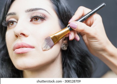 Beautiful Arabian Woman At Beauty Salon With A Nice Makeup. Make Up Artist Apply Foundation Cream On Face, Holding In Hands A Makeup Brushe On A Dark Or Black Background.