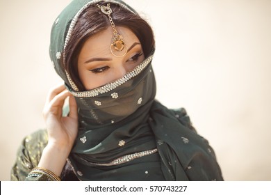 Beautiful Arabian woman portrait outdoors. Young Hindu woman. Portrait of beauty Indian model with bright make-up who hiding her face behind the veil standing over gold desert background.