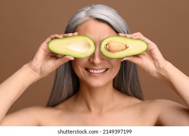 Beautiful Asian 50s Mature Grey-haired Woman Covering Eyes With Pieces Of Fresh Avocado Isolated On Brown. Attractive Mature Lady Recommends Oil-rich Foods. Healthy Eating And Lifestyle Concept