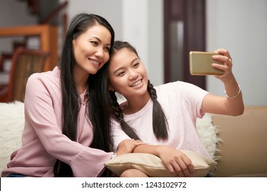 Beautiful Asian Adult Woman With Charming Teen Girl Sitting On Sofa And Taking Selfie With Phone