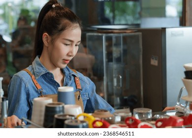 The beautiful Asian barista wears navy blue shirt and brown apron. Making coffee orders behind the coffee machine counter. front  beautifully arranged coffee cups. At small, minimalist cafe - Powered by Shutterstock