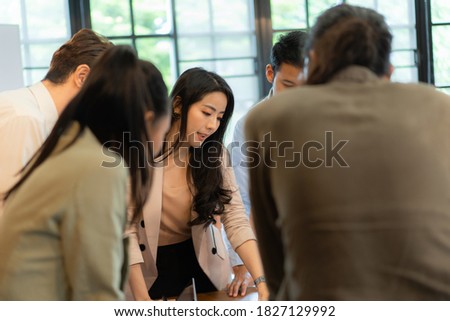 Beautiful Asian businesswoman in cream suit standing near meetingroom table with colleagues and explaining her new business plan ideas during meeting in the office meeting room.