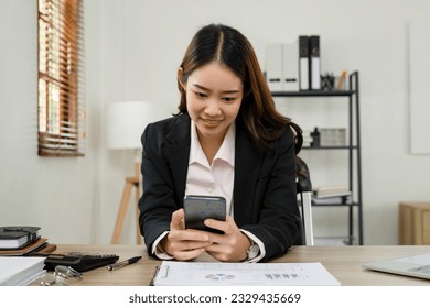 Beautiful asian businesswoman in formal black suit sitting at desk in workplace or office working on analyzing business data and charts, planning strategies and messaging and communicating with smartp
