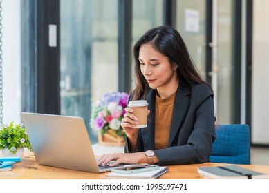 Beautiful Asian businesswoman holding coffee sitting in an office chair using a laptop keyboard.