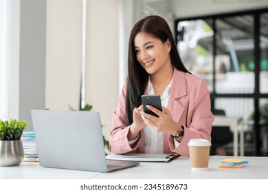 A beautiful Asian businesswoman looking at her laptop screen while using her smartphone at her desk in a modern office.