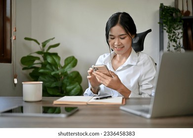 A beautiful Asian businesswoman texting with her friends or checking emails on her smartphone while sitting at her desk in her office.