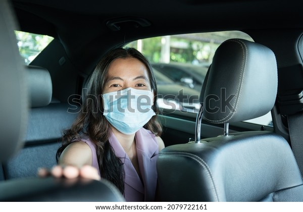Beautiful Asian businesswoman wear a hygiene\
protective face mask taking a back seat in taxi portrait, happy\
Asian woman passenger with face mask traveling by using taxi during\
the COVID-19\
pandemic.
