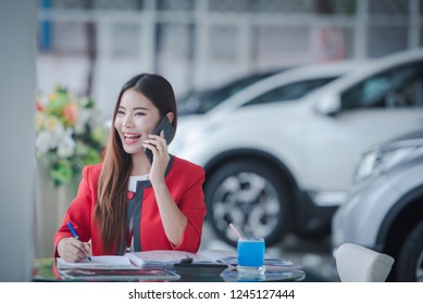 A beautiful Asian car saleswoman smiles happily while talking to a customer on the phone, having a calculator ready to calculate the price of a new car. - Powered by Shutterstock