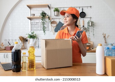 Beautiful Asian Delivery Woman In An Orange Uniform Send Package Products For Cook Customers In Kitchen Via Smartphone. Domestic And International Transport And Logistics