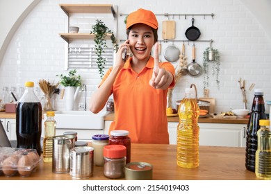 Beautiful Asian Delivery Woman In An Orange Uniform Send Package Products For Cook Customers In Kitchen Via Smartphone. Domestic And International Transport And Logistics