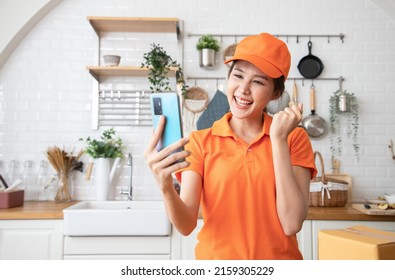 Beautiful Asian Delivery Woman In An Orange Uniform Send Package Products For Cook Customers In Kitchen Via Smartphone. Domestic And International Transport And Logistics