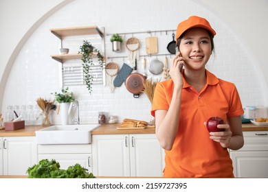 Beautiful Asian Delivery Woman In An Orange Uniform With Package Products For Cook Customers In Kitchen Via Smartphone. Domestic And International Transport And Logistics