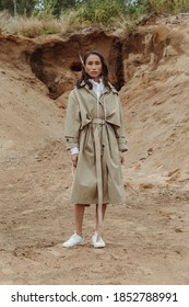 Beautiful Asian Fashionista In A Trench Coat Posing In Front Of Sand Pit. Vertical Image.