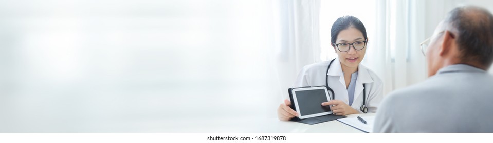 Beautiful Asian Female Doctor Talking Information About Virus To Elderly Patient With Digital Tablet. Physician Wear Glasses Giving Advice To Senior Man In Medical Room Of Hospital. Banner, Panoramic.