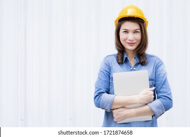 Beautiful Asian Female Engineer In Jeans Dress Wearing Yellow Safety Hard Hat Using Laptop Computer Notebook Doing Job At Construction Site Outside Office. Idea For Modern Working Woman.