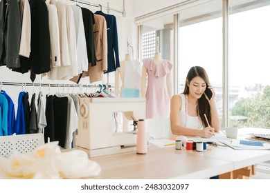 Beautiful Asian female fashion designers or dressmakers make phone calls with a customer for the new collection with samples of fabrics and sketches on paper. Fashion designer working in the studio. - Powered by Shutterstock