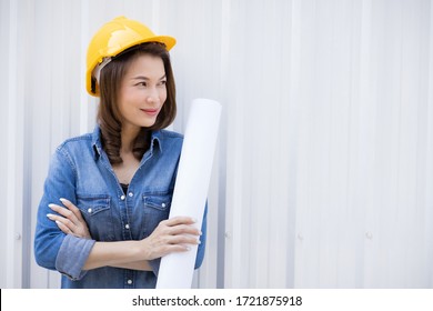 Beautiful Asian Female In Jeans Dress Wearing Yellow Safety Hat And Holding Roll Of White Paper Plan Standing In Front Of Metal Sheet Wall At Construction Site Outside Office. Idea For Working Woman.