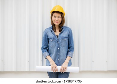 Beautiful Asian Female In Jeans Dress Wearing Yellow Safety Hat And Holding Roll Of White Paper Plan Standing In Front Of Metal Sheet Wall At Construction Site Outside Office. Idea For Working Woman.