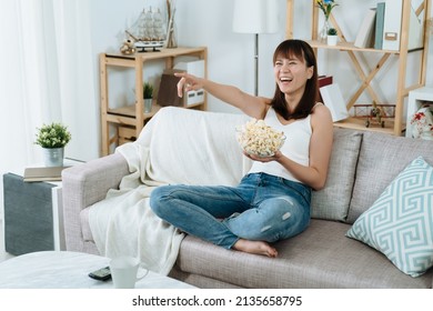 beautiful asian female sitting with popcorn on her sofa is watching a amusing show in her place. chinese woman is laughing and pointing at the screen for some funny jokes the tv host tells. 