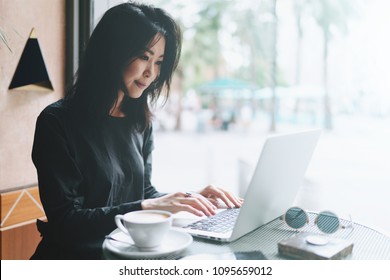 Beautiful Asian Female Student Sitting With Portable Laptop In Modern Coffee Shop. Successful Female Freelancer Using Portable Computer For A Distance Work During Morning Breakfast In Cafe