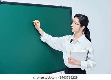 A beautiful Asian female teacher in white heating and glasses is holding a chalk in front of a green chalkboard and giving an Internet lecture with a confident expression and pose. - Powered by Shutterstock