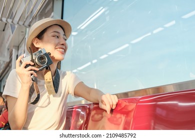 Beautiful Asian female tourist with face mask sits in a red seat, traveling by train, taking snapshot photo, transporting in suburb view, enjoy passenger lifestyle by railway, happy journey vacation.