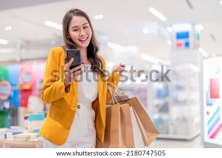 Beautiful asian female woman girl is holding shopping bags, using a smart phone and smiling checking discount promotion or coupon doing shopping in female boutique store shopping mall cheerful smiling