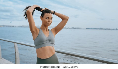 Beautiful, Asian fit girl in sportswear standing at embankment of sea makes ponytail looks at camera with confident face expression. Sporty American young woman training outdoors.