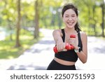 Beautiful asian fitness female sportswoman lifting dumbbell weight training after finish morning run outdoor. Happy smiling athlete woman excercising under the tree in the park while staring at camera