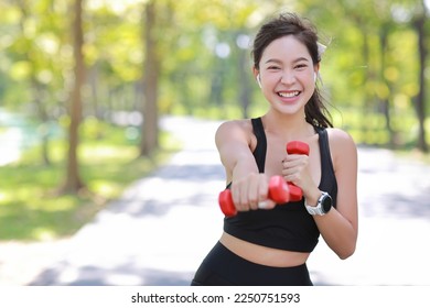 Beautiful asian fitness female sportswoman lifting dumbbell weight training after finish morning run outdoor. Happy smiling athlete woman excercising under the tree in the park while staring at camera