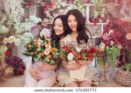 Beautiful asian florist girls making bouquet of flowers on table for sale against floral bokeh background in flower shop indoors. Two attractive asian females florists working in store. 2 playful