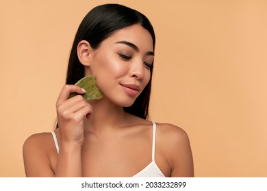 Beautiful Asian girl with beauty Gua Sha quartz stone for massage. Beige background isolated. Cosmetic natural trend. Traditional medicine and cosmetology concept.