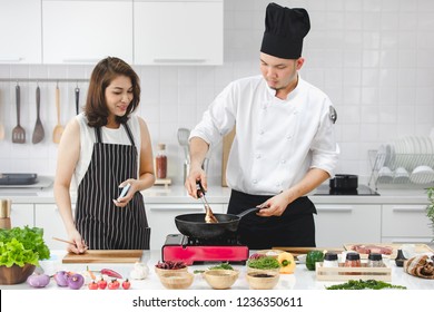 Beautiful Asian Girl In A Black Apron Smiles Happy In A Cooking Class Seeing The Chef Cook In A Saucepan To Make A Steak In A Modern, Clean Kitchen. Many Ingredients On The Table.