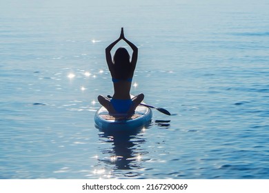 Beautiful asian girl in a blue bikini is engaged in water sports. She sits on a surf board and laughs. in the ocean against the background of sunset. Woman on paddle boarding  Sup.