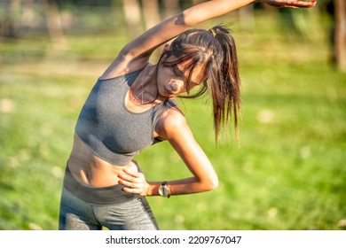 Beautiful Asian Girl Dressed In Sporty Clothes Stretching Her Body In A City Park.