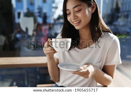Beautiful asian girl drinking coffee in cafe by the window, looking at cup of cappuccino and smiling on summer day.
