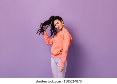 Beautiful Asian Girl In Earrings Standing On Purple Background. Studio Shot Of Charming Curly Chinese Woman Playing With Hair.