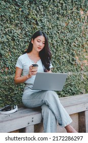 Beautiful Asian Girl Enjoying Coffee While Working Outdoors On A Laptop Connected To Public Wifi. Stylish College Student Studying Online While Spending Her Free Time In The Park.