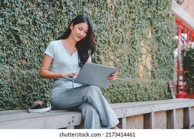 Beautiful Asian Girl Enjoying Coffee While Working Outdoors On A Laptop Connected To Public Wifi. Stylish College Student Studying Online While Spending Her Free Time In The Park.