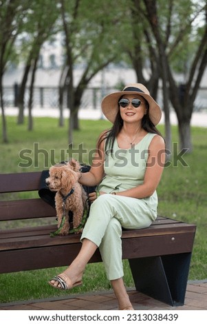 A beautiful Asian girl (Kazakh) with a hat and sunglasses sits on a bench in the park with her dog (mini poodle). Summer portrait of a young woman.