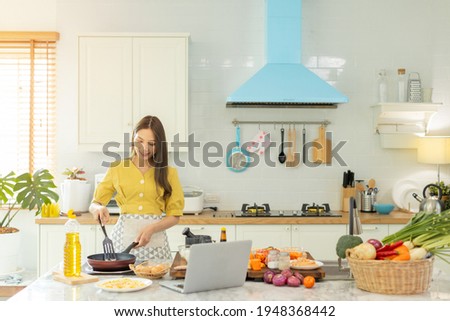 A beautiful Asian girl makes breakfast by looking at how to make it with her laptop computer with a happy face. At her own house