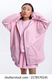 Beautiful Asian Girl In A Pink Casual Tracksuit Posing Against A White Wall In A Photo Studio