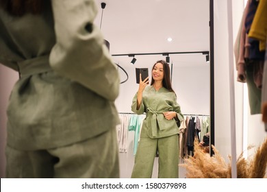 Beautiful Asian girl trying on stylish suit happily taking selfie in mirror in dressing room of modern clothes store