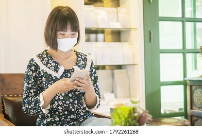 Beautiful Asian Girl Wearing Facemask Protection Contagious Coronavirus Covid-19, Using Smart Phone Mobile Online Internet Café Relaxing, Stay Safe Healthy Socializing In Public Area Social Distancing