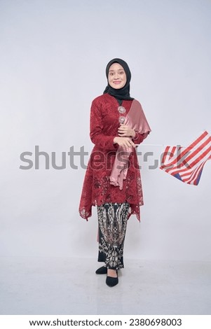 Beautiful Asian girl wearing traditional kebaya with hijab, happily holding Malaysian flag isolated over pink background. Independence day, new year nationalism concept.