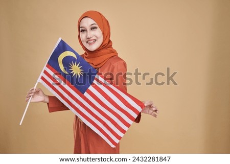 Beautiful Asian girl wearing traditional kebaya with hijab, happily holding Malaysian flag isolated over beige background. Independence day, new year nationalism concept.